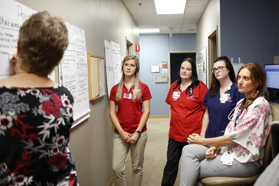 A group of nurses gather around to listen to a head nurse at a whiteboard.