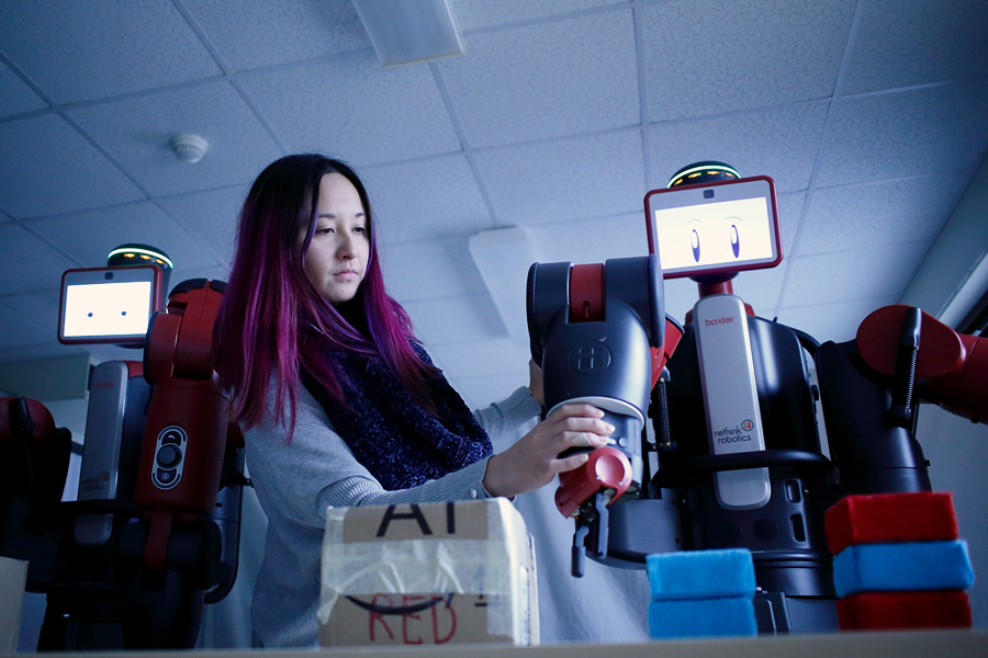 A student adjusts the arm of a robot. The robot is black, silver, and red, and the arm has an attachment for grabbing items.