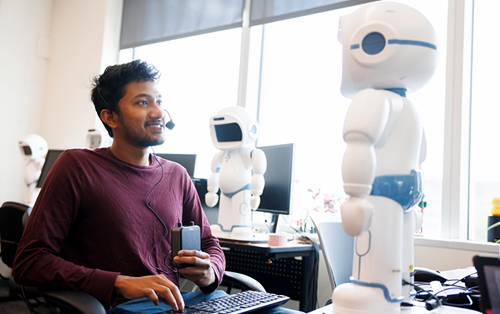 A student is seated with a keyboard and headset, facing a small, white, humanoid robot.