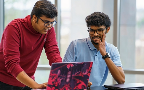 Two men discuss something they are looking at on a laptop.