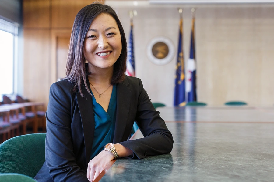 A woman in a suit smiles in a courtroom with flags in the background.