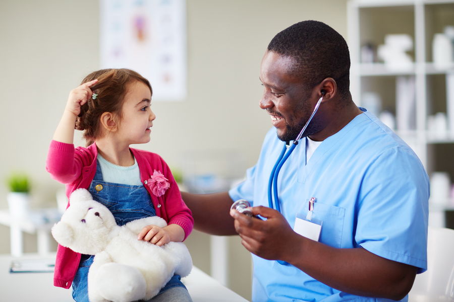 A man in scrubs holding a stethoscope up to a smiling girl holding a teddy bear