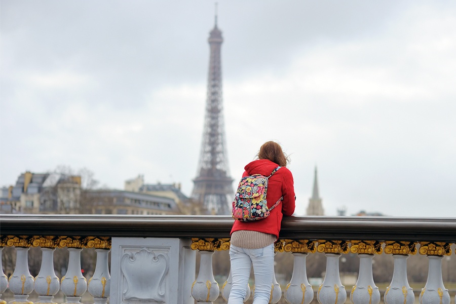 A student in a red jacket looks over a bridge at the Eiffel Tower.