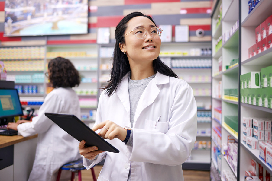 A pharmacy technician consults a tablet and looks at a shelf of patient medications.