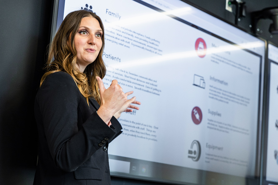 A woman in a suit gives a presentation and gestures to a large screen behind her.