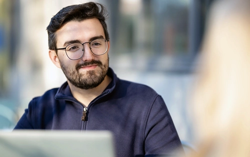 A working professional with laptop listens to a colleague.