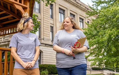 A woman holding a clipboard and stethoscope converses with a colleague outside a courthouse.