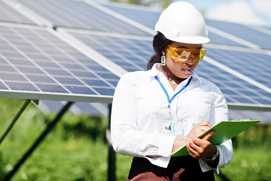 A person in a hard hat and protective eyewear stands in front of solar panels and makes notes on a clipboard.